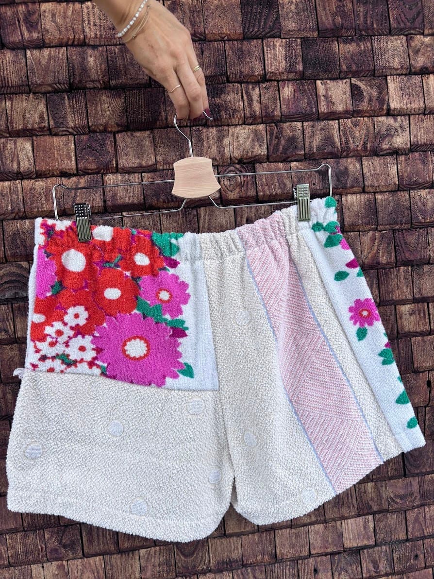 Rückansicht der Frottee-Patchwork-Shorts „Apricot '78“ von Old Wave an einem Kleiderbügel. Diese Seite präsentiert die asymmetrische Anordnung der upgecycelten Materialien: Das linke Hosenbein zeigt oben das auffällige, pink-rote Retro-Blumenmuster auf weißem Grund über einer hellbeigen Frotteefläche. Das rechte Hosenbein besteht primär aus dem hellbeigen Strukturstoff und wird an der Außenkante von einer feinen weißen Stoffbahn mit pinken Blütenmotiven flankiert.