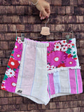 Upcycling-Shorts „Apricot '78“ von Old Wave auf einem Kleiderbügel vor einer rustikalen Holzwand. Das lebhafte Patchwork-Design aus Frottee-Handtüchern zeigt auf der linken Seite ein buntes Vintage-Blumenmuster in Pink, Rot und Weiß, kombiniert mit einer strukturierten, hellbeigen Mittelfläche. Das rechte Hosenbein kombiniert oben ebenfalls das florale Muster mit einer Fransenborte und geht unten in ein vertikal gestreiftes Muster in Weiß, Rosa und Grün über.