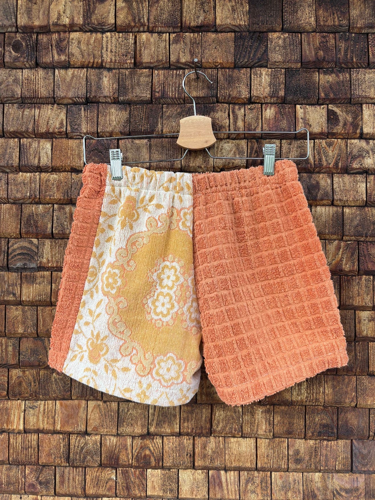 Rückansicht der Upcycling-Frotteeshorts „Apricot Dream ‘69“ von Old Wave auf einem Kleiderbügel vor einer Holzwand. Die Rückseite zeigt ein harmonisches Muster-Layout: Die linke Gesäßhälfte setzt das kunstvolle, gelb-weiße Ornament- und Fliesenmuster fort, umrahmt von einem apricotfarbenen Seitenstreifen. Die rechte Gesäßhälfte besteht vollständig aus einem strukturierten, unifarbenen Frotteestoff in warmem Apricot.