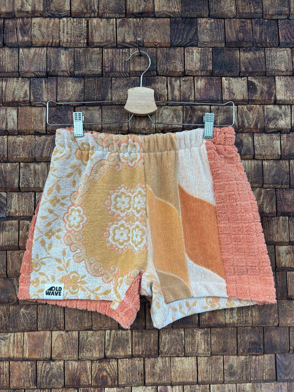 Vorderansicht der Upcycling-Frotteeshorts „Apricot Dream ‘69“ von Old Wave auf einem Kleiderbügel vor einer rustikalen Holzwand. Das filigrane Vintage-Design kombiniert ein rechtes Hosenbein mit gelb-weißen Retro-Mustern und Paisley-Ornamenten samt Logo-Patch am Saum mit einem linken Hosenbein, das ein abstrakt geschwungenes Wellenmuster in Apricot-, Creme- und Beigetönen zeigt, flankiert von einem melierten apricotfarbenen Außenstreifen.