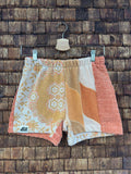Vorderansicht der Upcycling-Frotteeshorts „Apricot Dream ‘69“ von Old Wave auf einem Kleiderbügel vor einer rustikalen Holzwand. Das filigrane Vintage-Design kombiniert ein rechtes Hosenbein mit gelb-weißen Retro-Mustern und Paisley-Ornamenten samt Logo-Patch am Saum mit einem linken Hosenbein, das ein abstrakt geschwungenes Wellenmuster in Apricot-, Creme- und Beigetönen zeigt, flankiert von einem melierten apricotfarbenen Außenstreifen.