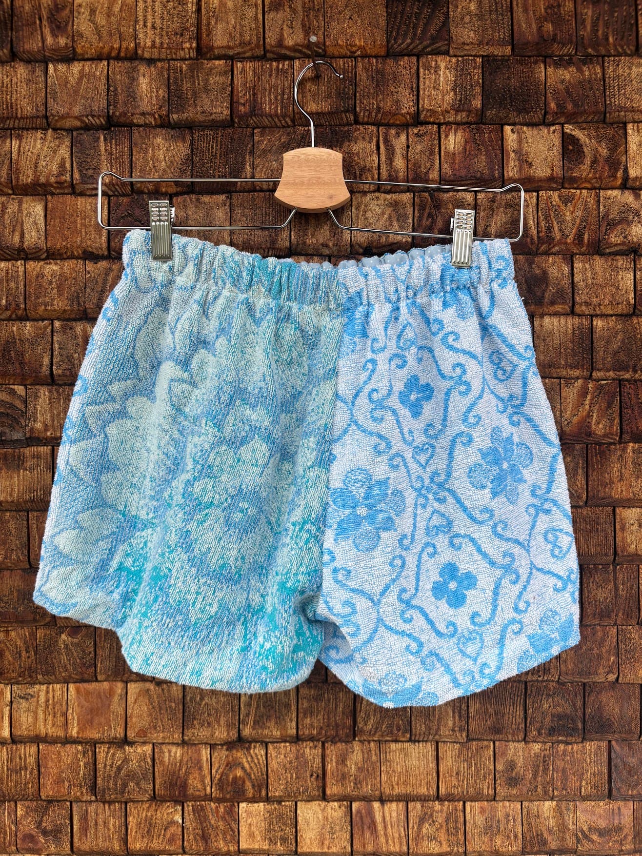 Rückansicht der Upcycling-Frotteeshorts „Aqua Waves ‘82“ von Old Wave auf einem Kleiderbügel vor einer Holzwand. Die Rückseite führt das harmonische Farbblock-Konzept in kühlen Wassertönen fort: Die linke Gesäßhälfte zeigt ein großes, rundes Mandala- und Wellenornament in Hellblau und Türkis, während die rechte Gesäßhälfte komplett mit dem blau-weißen Vintage-Fliesenmuster gestaltet ist.