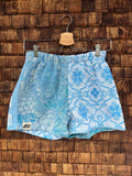 Vorderansicht der Upcycling-Frotteeshorts „Aqua Waves ‘82“ von Old Wave auf einem Kleiderbügel vor einer rustikalen Holzwand. Das maritime Patchwork-Design besticht durch eine exakt hälftige Teilung: Das linke Hosenbein zeigt ein hellblau-türkis schattiertes, florales Muster mit Wellen-Vibe und dem Old Wave-Logo am Saum, während das rechte Hosenbein ein kontrastierendes, blau-weißes Ornamentmuster im mediterranen Fliesenstil präsentiert.