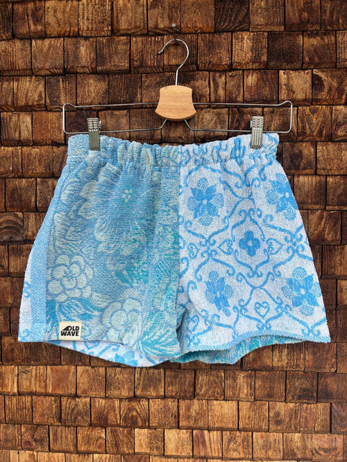 Vorderansicht der Upcycling-Frotteeshorts „Aqua Waves ‘82“ von Old Wave auf einem Kleiderbügel vor einer rustikalen Holzwand. Das maritime Patchwork-Design besticht durch eine exakt hälftige Teilung: Das linke Hosenbein zeigt ein hellblau-türkis schattiertes, florales Muster mit Wellen-Vibe und dem Old Wave-Logo am Saum, während das rechte Hosenbein ein kontrastierendes, blau-weißes Ornamentmuster im mediterranen Fliesenstil präsentiert.