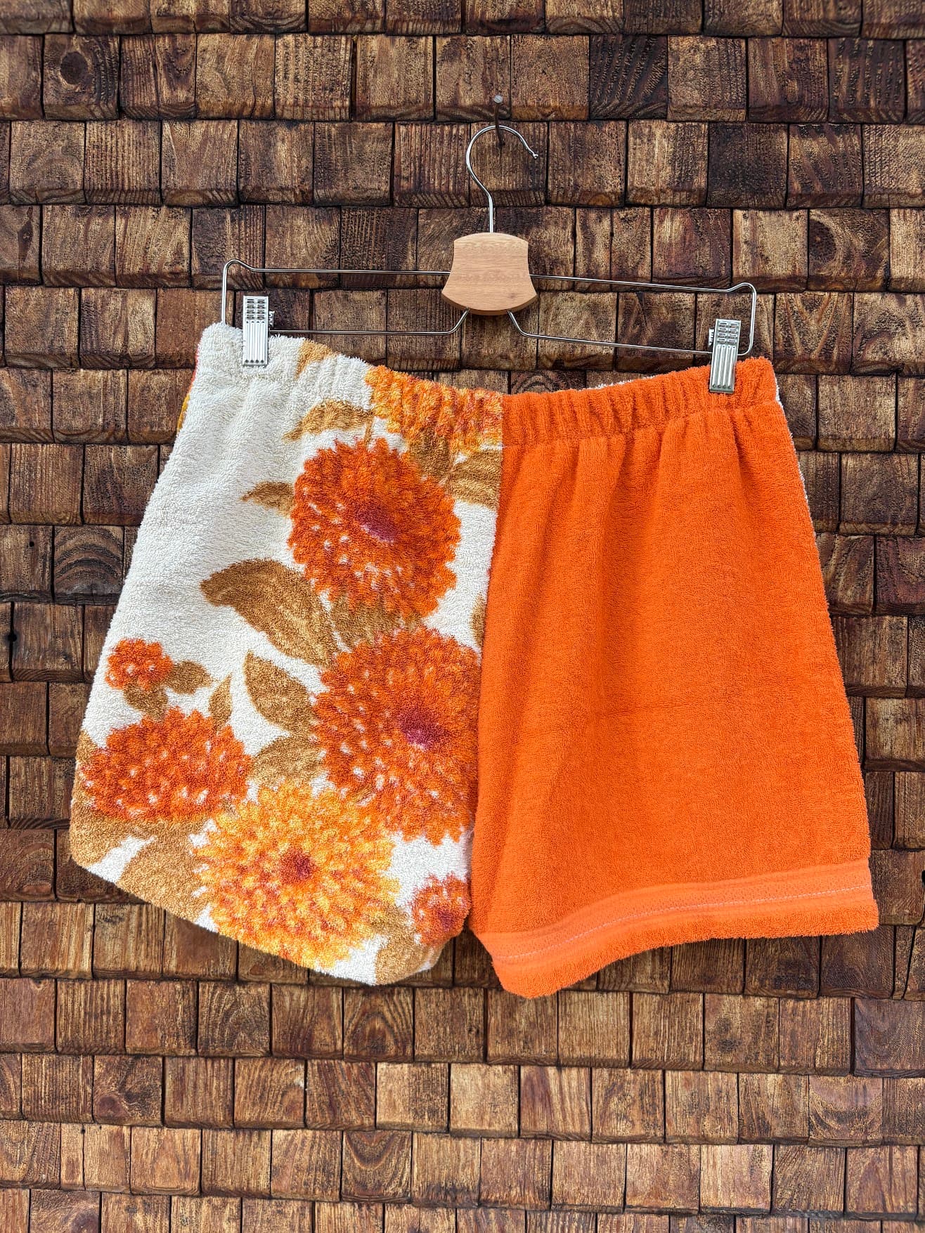 Rückansicht der Upcycling-Frotteeshorts „Autumn Bloom ‘72“ von Old Wave auf einem Kleiderbügel vor einer Holzwand. Die Rückseite spiegelt das asymmetrische Design wider: Die linke Gesäßhälfte zeigt großflächige, nostalgische Retro-Blüten in Orange auf weißem Grund, während die rechte Gesäßhälfte komplett aus weichem, orangefarbenem Frotteestoff mit einem farblich abgesetzten Saum besteht.