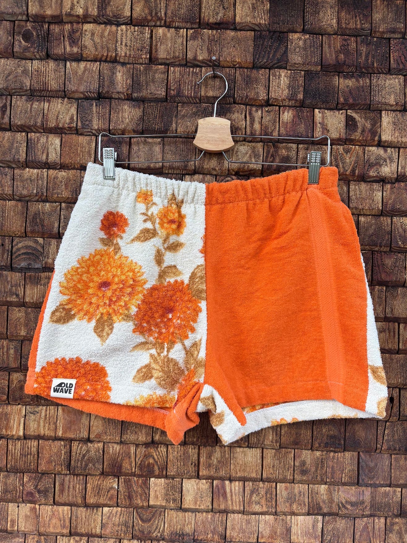 Vorderansicht der Upcycling-Frotteeshorts „Autumn Bloom ‘72“ von Old Wave auf einem Kleiderbügel vor einer rustikalen Holzwand. Das herbstliche 70er-Jahre-Retro-Design ist zweigeteilt: Das rechte Hosenbein besteht aus weißem Frottee mit einem filigranen Blumenmuster in warmen Orange- und Brauntönen sowie dem Logo-Patch am Saum. Das linke Hosenbein ist in kräftigem, unifarbenem Orange gehalten und mit hellen Kontraststreifen an den Seiten versehen.