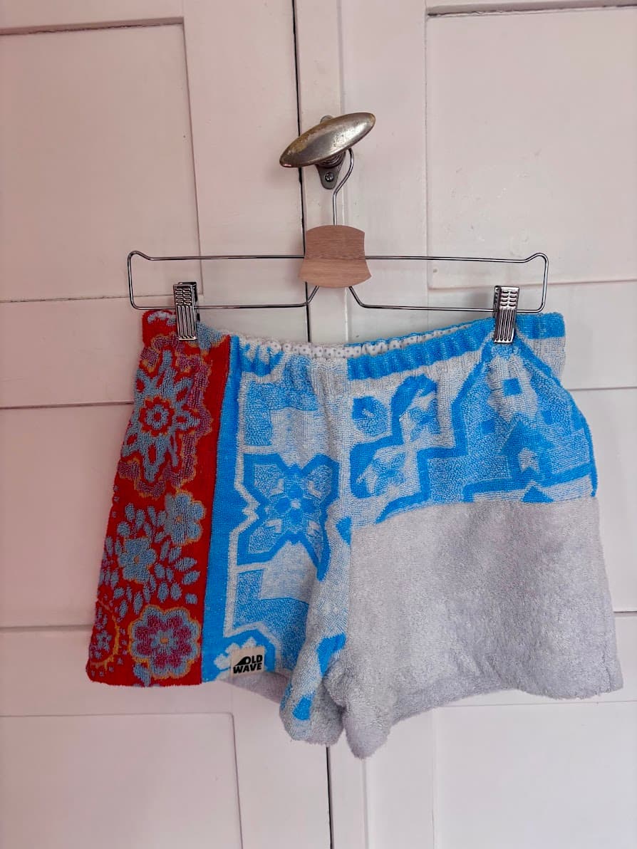 Frottee-Upcycling-Shorts „Azur Coast 65“ von Old Wave auf einem Kleiderbügel vor einer weißen Holztür. Zu sehen ist das dreigeteilte Patchwork-Design der Vorderseite: Ein hellgraues Hosenbein trifft auf ein geteiltes Hosenbein mit azurblauem Ornamentmuster oben und hellgrauem Stoff unten, ergänzt durch einen seitlichen Streifen aus rotem Vintage-Handtuchstoff.
