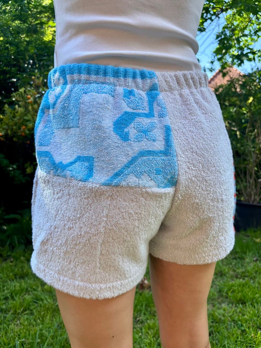 Rückansicht von schräg hinten links der „Azur Coast 65“ Frottee-Shorts am Model. Das Design kombiniert eine schlichte weiße Frotteefläche am Gesäß mit einem azurblau-weißen Kachelmuster im oberen Bereich des linken Hosenbeins.