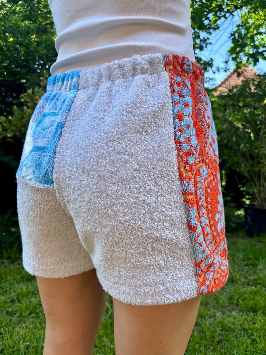Trageansicht der Upcycling-Shorts „Azur Coast 65“ von Old Wave von schräg hinten rechts im Garten. Zu sehen ist das weiße Frottee-Gesäßdesign, das am rechten Bein in eine auffällige Patchwork-Bahn aus rotem Vintage-Handtuchstoff mit hellblauen Ornament-Verzierungen übergeht.