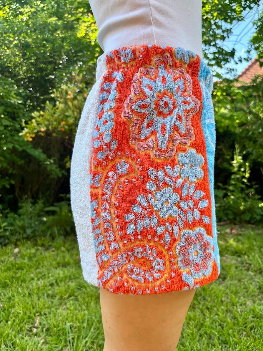 Linke Seitenansicht der upgecycelten Shorts „Azur Coast 65“ von Old Wave im Freien. Das Bild fokussiert den kontrastierenden, leuchtend roten Handtuchstoff, welcher mit großen Retro-Blumenmotiven und barocken Paisley-Formen in Hellblau und Orange gemustert ist.
