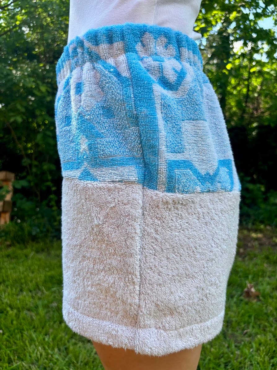 Rechte Seitenansicht der Frottee-Patchwork-Shorts „Azur Coast 65“ vor grünem Gartenhintergrund. Diese Perspektive zeigt die vertikale Stoffteilung: Oben ein azurblaues mediterranes Fliesenmuster auf weißem Grund und unten schlichter, weißer Frotteestoff.