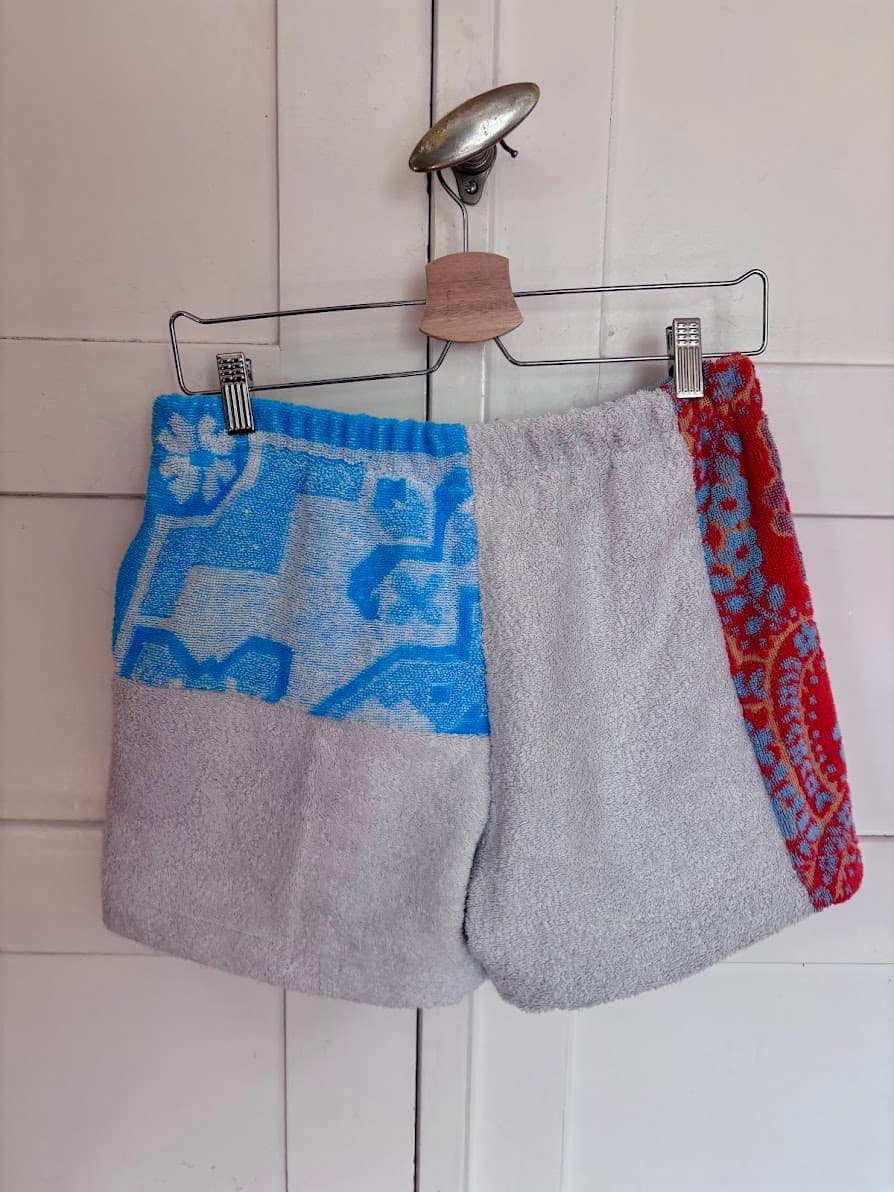 Rückansicht der „Azur Coast 65“ Frottee-Shorts von Old Wave an einem Kleiderbügel. Diese Perspektive zeigt die Rückseite der Shorts mit einer Kombination aus leuchtend rotem Handtuchstoff samt weiß-orangen Retro-Mustern auf der linken Seite, einer mittleren Partie im azurblau-weißen Fliesen-Look und einem hellgrauen Frotteeelement unten rechts.