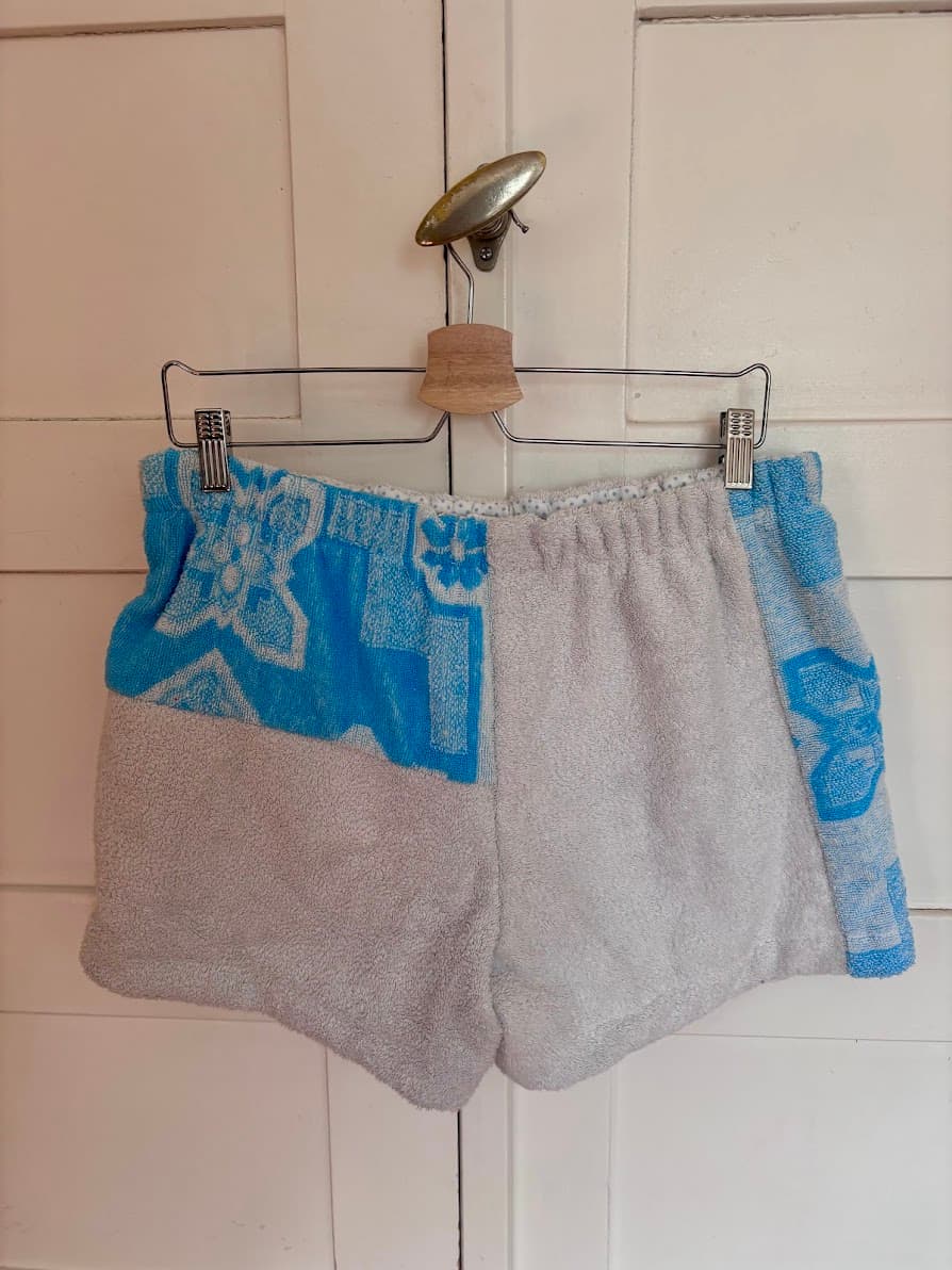 Rückansicht der Frottee-Patchwork-Shorts „Azure di Portofino 64“ von Old Wave an einem Kleiderbügel. Diese Seite präsentiert den umgekehrten Materialmix der upgecycelten Handtücher: Ein vorwiegend hellgraues Gesäßdesign, das auf der einen Hosenbeinseite im unteren Bereich in das markante azurblau-weiße Fliesen- und Retromuster übergeht.