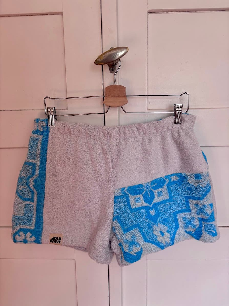 Upcycling-Shorts „Azure di Portofino 64“ von Old Wave auf einem Kleiderbügel vor einer weißen Holzwand. Das asymmetrische Design aus Frottee-Handtüchern kombiniert ein schlichtes hellgraues Hosenbein mit einer geteilten Optik auf der anderen Seite, die oben ein leuchtend azurblaues Ornament- und Kachelmuster und unten ebenfalls hellgrauen Frotteestoff zeigt.