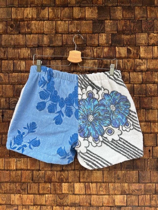 Rückansicht der Upcycling-Frotteeshorts „Blue Garden ‘75“ von Old Wave auf einem Kleiderbügel vor einer Holzwand. Die Rückseite spiegelt das asymmetrische Blumendesign wider: Auf der linken Gesäßhälfte setzt sich die hellblaue Optik mit den dunkelblauen Blütenranken fort, während die rechte Gesäßhälfte vollständig mit dem ausdrucksstarken, schwarz-weiß-blauen Retro-Blumenmuster gestaltet ist.
