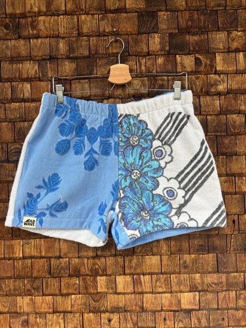 Vorderansicht der Upcycling-Frotteeshorts „Blue Garden ‘75“ von Old Wave auf einem Kleiderbügel vor einer rustikalen Holzwand. Das kontrastreiche Patchwork-Design besticht durch eine zweigeteilte Vorderseite: Das linke Hosenbein besteht aus einem unifarbenen, hellblauen Frotteestoff mit dunkelblauen Vintage-Blumenranken und dem Old Wave-Logo am Saum. Das rechte Hosenbein zeigt große, leuchtend blaue Blüten mit grafischen, schwarz-weißen Streifenelementen im Retro-Stil.