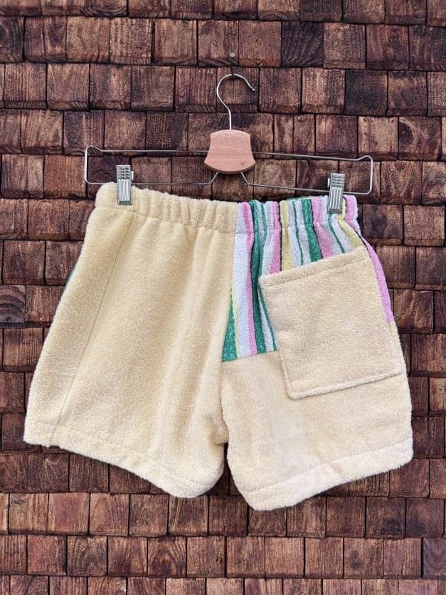 Rückansicht der Upcycling-Frotteeshorts „Candy Stripe ‘95“ von Old Wave auf einem Kleiderbügel vor einer Holzwand. Die Rückseite präsentiert eine unifarbene linke Gesäßhälfte aus weichem, beigegelbem Vintage-Frottee. Die rechte Seite führt das vertikale Streifenmuster am Bund fort, läuft unten hellgelb aus und ist mit einer großen, aufgesetzten Gesäßtasche im passenden beigegelben Frotteestoff versehen.