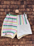 Vorderansicht der Upcycling-Frotteeshorts „Candy Stripe ‘95“ von Old Wave auf einem Kleiderbügel vor einer rustikalen Holzwand. Das farbenfrohe Streifendesign zeigt ein linkes Hosenbein mit horizontalen Retro-Streifen in Pink, Grün und Gelb auf weißem Grund samt dem Old Wave-Logo am Saum. Das rechte Hosenbein kombiniert vertikale Streifen im gleichen Muster am Bund mit einem unteren Teil aus schlichtem, hellgrauem Frotteestoff.