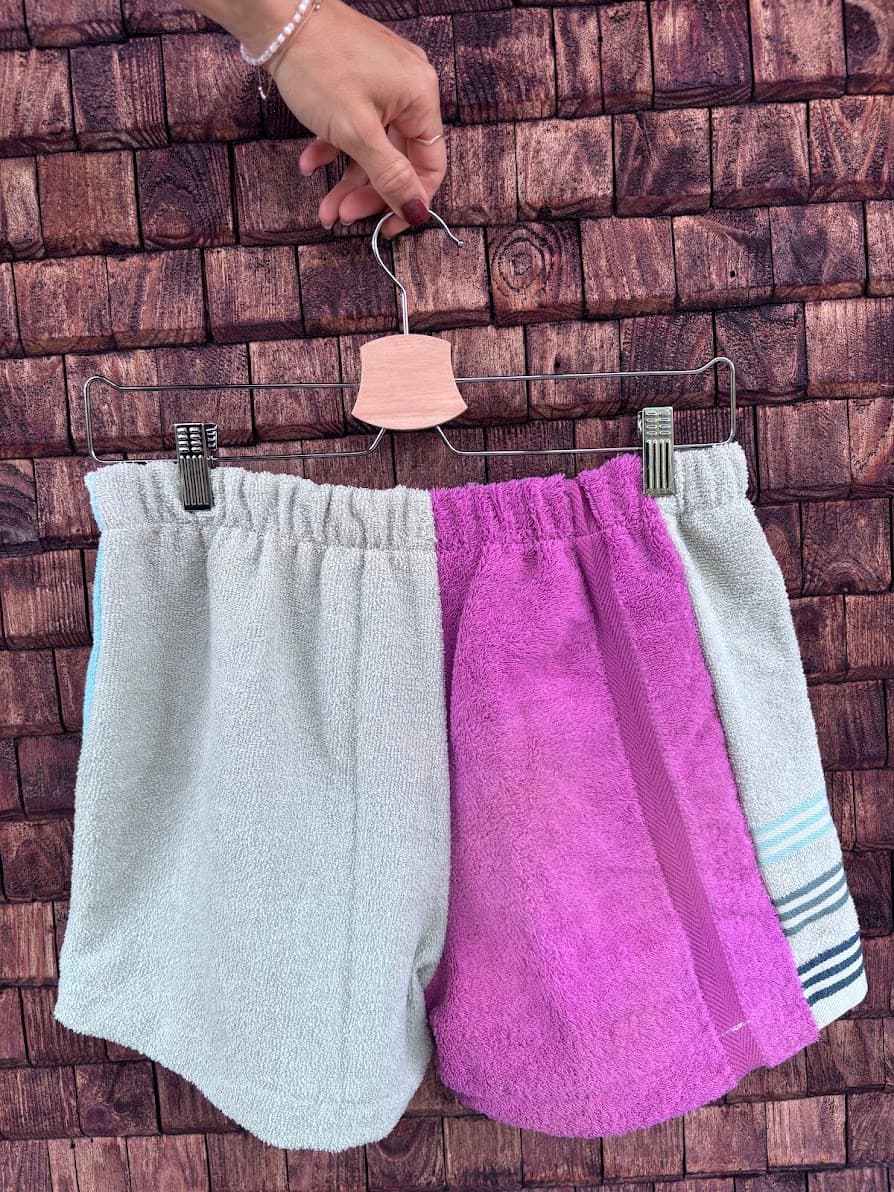 Rückansicht der Frottee-Patchwork-Shorts „Capri '72“ von Old Wave an einem Kleiderbügel. Diese Perspektive zeigt die asymmetrische Gesäßpartie aus upgecycelten Handtüchern: Das linke Hosenbein besteht vorwiegend aus einem kräftigen lila-magentafarbenen Frotteestoff, während das rechte Hosenbein in sanftem Mint gehalten ist und am äußeren Rand dezent das blau-weiß gestreifte Streifenmuster aufgreift.