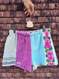 Upcycling-Shorts „Capri '72“ von Old Wave auf einem Kleiderbügel vor einer rustikalen Holzwand. Das farbenfrohe Frottee-Patchwork-Design zeigt auf der linken Seite ein mintfarbenes Hosenbein, das im unteren Drittel mit einem blau-weiß gestreiften Vintage-Muster abschließt. Die Mitte und das rechte Hosenbein kombinieren ein kräftiges Lila-Magenta mit einer hellblauen Stoffbahn, die an der Außenseite von einer weißen Bordüre mit leuchtend pinken Blumen verziert ist.