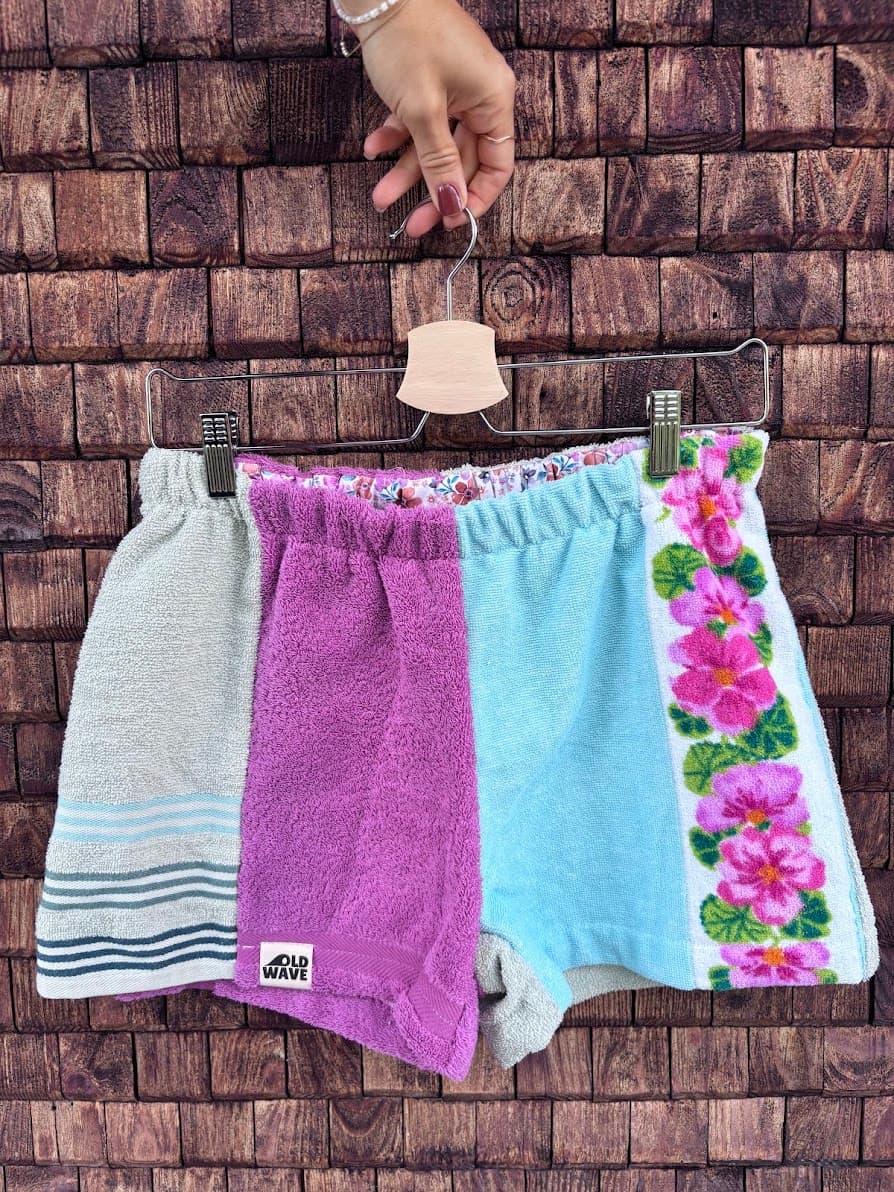 Upcycling-Shorts „Capri '72“ von Old Wave auf einem Kleiderbügel vor einer rustikalen Holzwand. Das farbenfrohe Frottee-Patchwork-Design zeigt auf der linken Seite ein mintfarbenes Hosenbein, das im unteren Drittel mit einem blau-weiß gestreiften Vintage-Muster abschließt. Die Mitte und das rechte Hosenbein kombinieren ein kräftiges Lila-Magenta mit einer hellblauen Stoffbahn, die an der Außenseite von einer weißen Bordüre mit leuchtend pinken Blumen verziert ist.