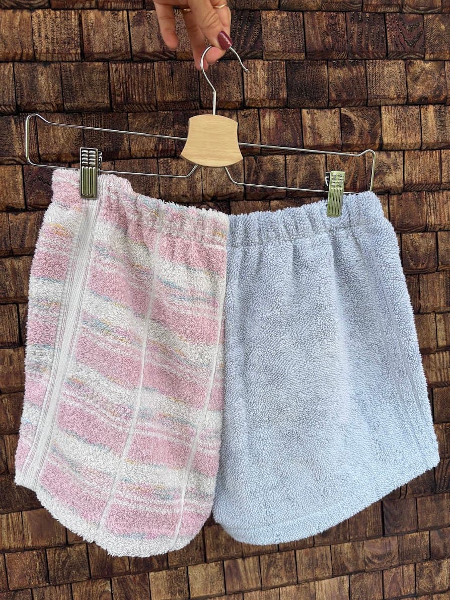 Rückansicht der Frottee-Patchwork-Shorts „Cherry '69“ von Old Wave an einem Kleiderbügel. Diese Seite zeigt die gespiegelte Anordnung der upgecycelten Vintage-Handtücher: Die linke Gesäßseite besteht aus dem weiß-rosa karierten Muster mit feinen bunten Streifenakzenten, während die rechte Seite in schlichtem Hellblau gehalten ist.