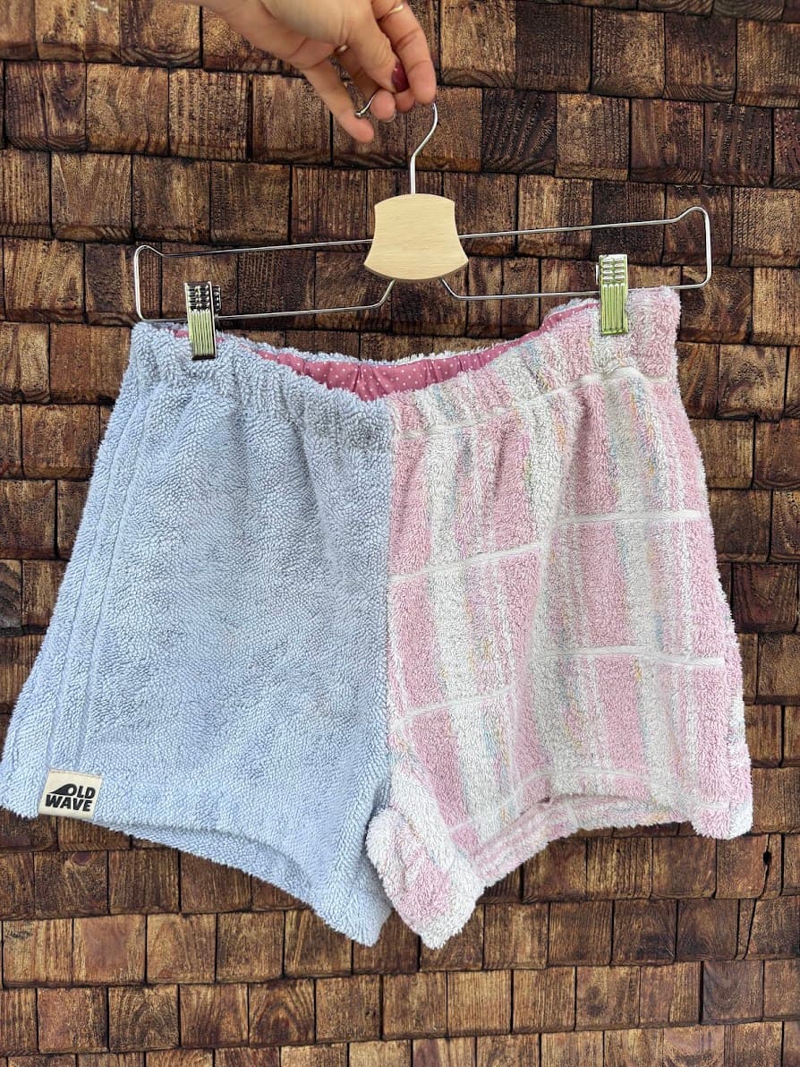 Upcycling-Shorts „Cherry '69“ von Old Wave auf einem Kleiderbügel vor einer rustikalen Holzwand präsentiert. Das Patchwork-Design aus weichem Frottee kombiniert ein schlichtes, hellblaues Hosenbein auf der linken Seite mit einem kontrastierenden rechten Hosenbein aus weiß-rosa kariertem Handtuchstoff.