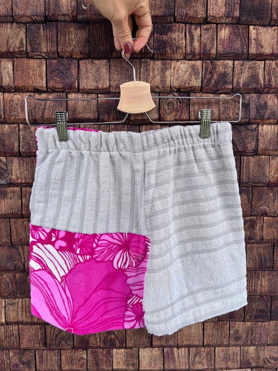 Rückansicht der Frottee-Patchwork-Shorts „Coral '84“ von Old Wave an einem Kleiderbügel. Diese Perspektive zeigt das asymmetrische Gesäßdesign aus upgecycelten Handtüchern: Die Rückseite besteht vorwiegend aus einem hellgrau strukturierten Frotteestoff, wobei das linke Hosenbein im unteren Drittel mit dem kontrastierenden, intensiv pink-weißen Vintage-Blumenmuster abschließt.