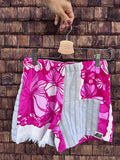 Upcycling-Shorts „Coral '84“ von Old Wave auf einem Kleiderbügel vor einer dunklen Holzwand. Das lebendige Patchwork-Design aus Frottee-Handtüchern kombiniert ein linkes Hosenbein in leuchtendem Pink mit weißen Südsee-Blumenmotiven und Fransensaum mit einem rechten Hosenbein aus hellgrauem Frotteestoff, das im oberen Bereich ebenfalls das pinke Blütenmuster aufgreift.