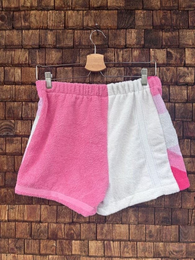 Rückansicht der Upcycling-Frotteeshorts „Coral Chevron ‘102“ von Old Wave auf einem Kleiderbügel vor einer Holzwand. Die Rückseite zeigt ein zweigeteiltes Patchwork-Design mit einer unifarbenen, kräftig rosafarbenen linken Gesäßhälfte und einer schlichten, weißen rechten Stoffhälfte aus weichem Vintage-Handtuchmaterial.