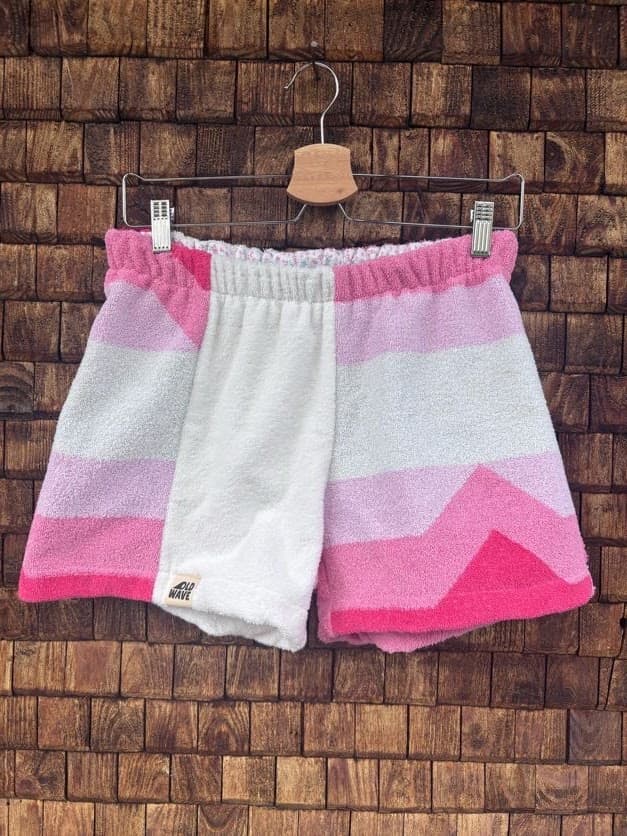 Vorderansicht der Upcycling-Frotteeshorts „Coral Chevron ‘102“ von Old Wave auf einem Kleiderbügel vor einer rustikalen Holzwand. Das asymmetrische Design kombiniert ein rechtes Hosenbein aus vorwiegend weißem Vintage-Frottee mit rosafarbenen Akzenten am Bund und Saum sowie ein linkes Hosenbein mit einem markanten, mehrfarbigen Chevron-Zickzack-Muster in verschiedenen Rosa- und Hellpinktönen.