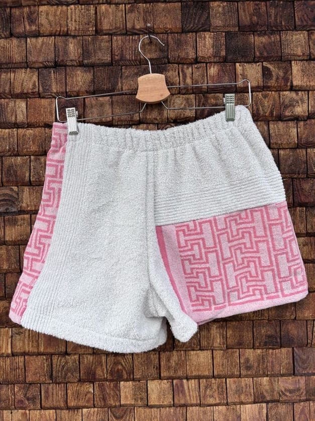 Rückansicht der Upcycling-Frotteeshorts „Coral Maze ‘86“ von Old Wave auf einem Kleiderbügel vor einer Holzwand. Die Rückseite spiegelt das asymmetrische Design wider: Die Basis ist in schlichtem Weiß gehalten, wobei die rechte Gesäßhälfte mit einem auffälligen korallenrosa Muster-Panel versehen ist und die linke Außenseite mit dem vertikalen, korallenrosa Zierstreifen abschließt.