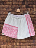 Vorderansicht der Upcycling-Frotteeshorts „Coral Maze ‘86“ von Old Wave auf einem Kleiderbügel vor einer rustikalen Holzwand. Die Hosenbasis besteht aus hellem, weißem Vintage-Frottee. Das linke Hosenbein zeigt im unteren Bereich ein korallenrosa Patchwork-Element mit einem geometrischen Labyrinthtmuster sowie das Old Wave-Logo am Saum, während das rechte Bein durch einen vertikalen korallenrosa Musterstreifen an der Außenseite ergänzt wird.
