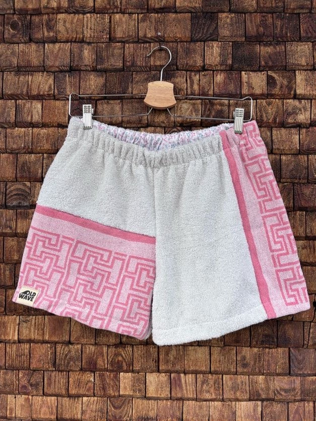 Vorderansicht der Upcycling-Frotteeshorts „Coral Maze ‘86“ von Old Wave auf einem Kleiderbügel vor einer rustikalen Holzwand. Die Hosenbasis besteht aus hellem, weißem Vintage-Frottee. Das linke Hosenbein zeigt im unteren Bereich ein korallenrosa Patchwork-Element mit einem geometrischen Labyrinthtmuster sowie das Old Wave-Logo am Saum, während das rechte Bein durch einen vertikalen korallenrosa Musterstreifen an der Außenseite ergänzt wird.