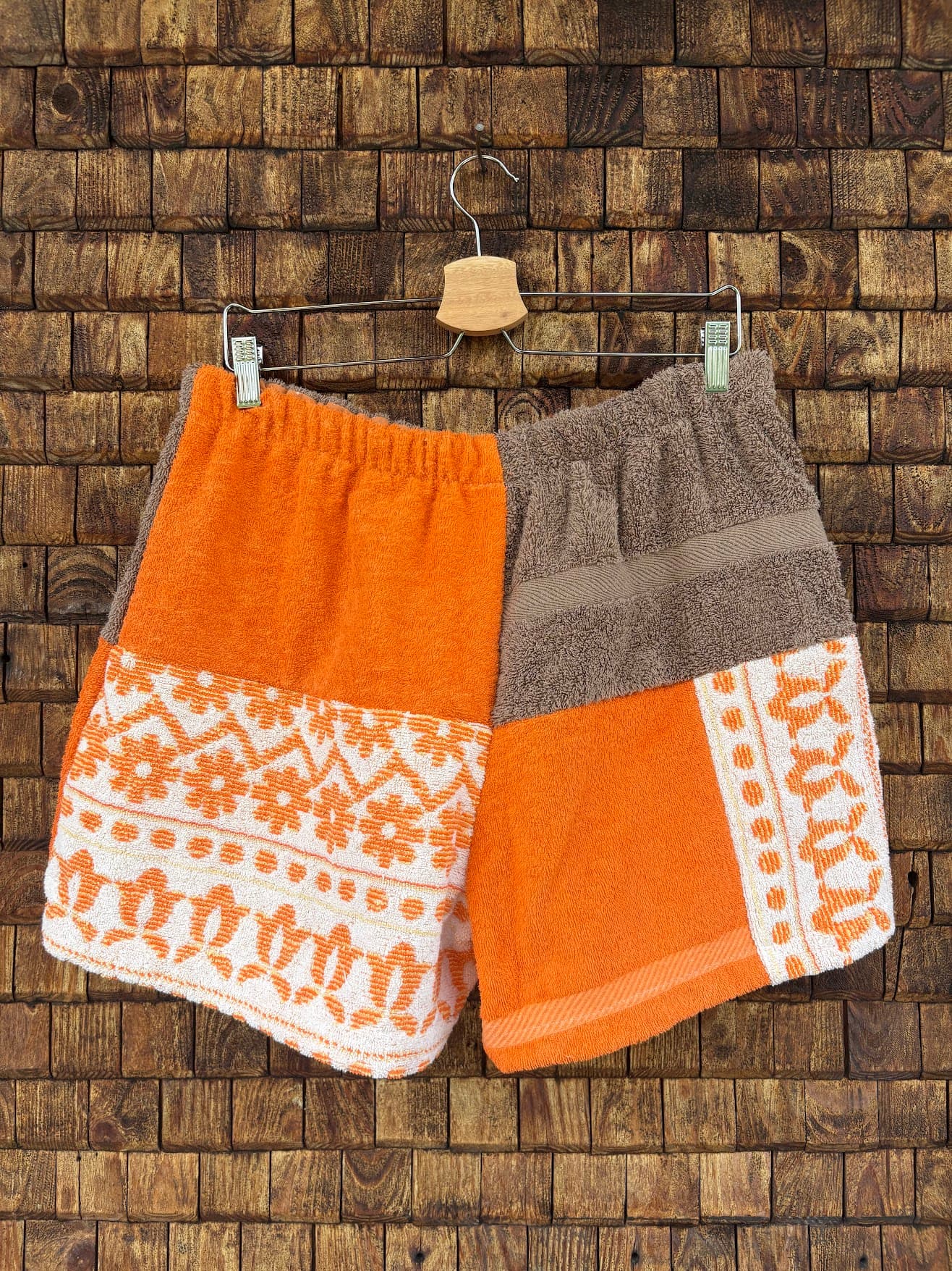 Rückansicht der Upcycling-Frotteeshorts „Coral Mosaic ‘76“ von Old Wave auf einem Kleiderbügel vor einer Holzwand. Die Rückseite spiegelt das vielseitige Color-Blocking-Konzept wider: Die linke Gesäßhälfte kombiniert ein oberes Panel in kräftigem Orange mit dem weiß-orangefarbenen Vintage-Mosaikmuster im unteren Bereich. Die rechte Gesäßhälfte zeigt oben braunes Frottee und unten oranges Frottee, welches an der Außennaht durch einen vertikalen Mosaik-Musterstreifen akzentuiert wird.