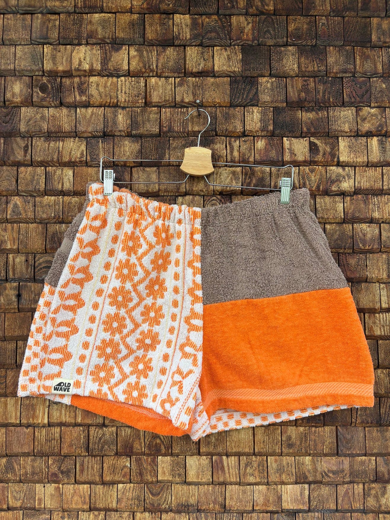 Vorderansicht der Upcycling-Frotteeshorts „Coral Mosaic ‘76“ von Old Wave auf einem Kleiderbügel vor einer rustikalen Holzwand. Das kreative Patchwork-Design zeigt eine asymmetrische Zweiteilung: Das rechte Hosenbein kombiniert eine obere Hälfte aus unifarbenem, braunem Frottee mit einer unteren Hälfte in leuchtendem Orange. Das linke Hosenbein besticht durch ein durchgehendes, weiß-orangefarbenes Retro-Mosaikmuster mit floralen Geometrie-Elementen und trägt das dezente Logo-Patch am Saum.