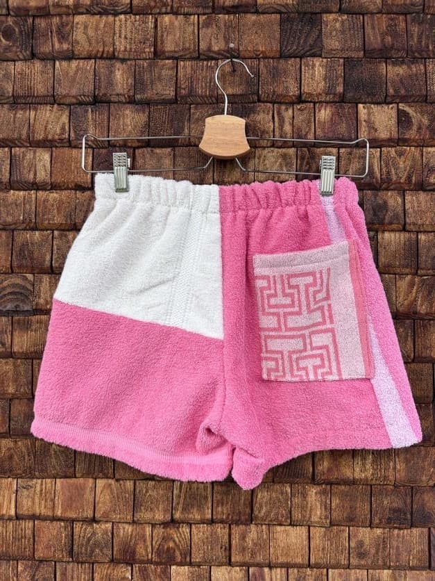 Rückansicht der Upcycling-Frotteeshorts „Coral Step ‘91“ von Old Wave auf einem Kleiderbügel vor einer Holzwand. Die asymmetrische Rückseite zeigt eine linke Gesäßhälfte, die zweigeteilt ist in einen weißen oberen und einen pinken unteren Bereich. Die rechte Hälfte ist größtenteils im kräftigen Pink gehalten, wird durch einen weißen Seitenstreifen an der Kante ergänzt und verfügt über eine aufgesetzte, hellrosa gemusterte Tasche.