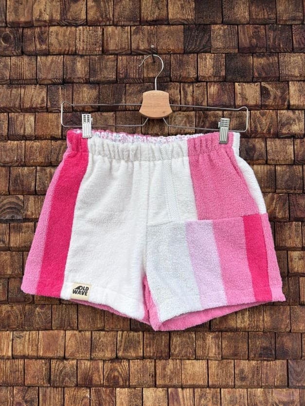 Vorderansicht der Upcycling-Frotteeshorts „Coral Step ‘91“ von Old Wave auf einem Kleiderbügel vor einer rustikalen Holzwand. Das auffällige asymmetrische Patchwork-Design kombiniert ein vorwiegend weißes linkes Hosenbein mit einem kräftig pinken Seitenstreifen an der Außenkante und dem Old Wave-Logo am Saum. Das rechte Hosenbein besteht im oberen Bereich aus pinkfarbenem Frottee und geht im unteren Teil in ein vertikales Streifenmuster in Weiß, Hellrosa und Pink über.