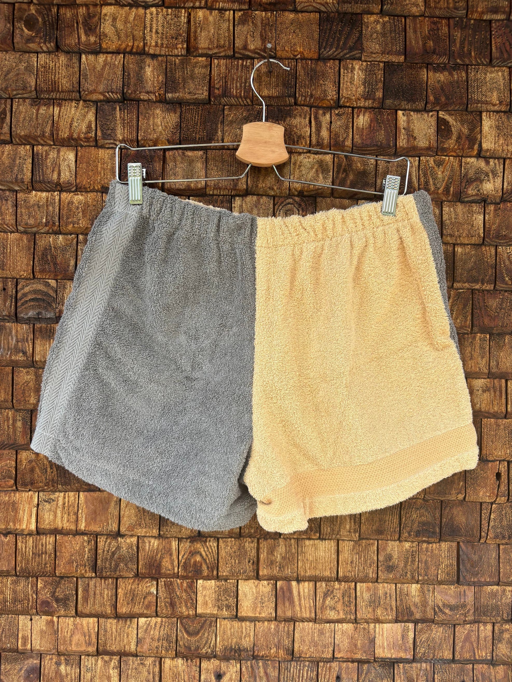 Rückansicht der Upcycling-Frotteeshorts „Cozy Grey & Sand ‘82“ von Old Wave auf einem Kleiderbügel vor einer Holzwand. Die Rückseite führt das harmonische Farbblock-Design fort, mit einer linken Gesäßhälfte in meliertem Grau, die an der Außenkante durch einen hellgrauen Streifen akzentuiert wird, und einer rechten Gesäßhälfte aus unifarbenem Frottee in warmem Sandgelb.