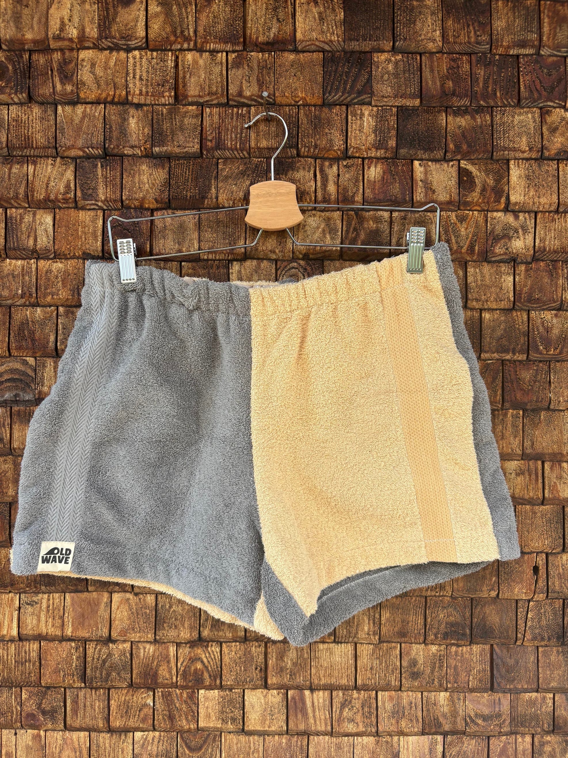 Vorderansicht der Upcycling-Frotteeshorts „Cozy Grey & Sand ‘82“ von Old Wave auf einem Kleiderbügel vor einer rustikalen Holzwand. Das minimalistische Two-Tone-Design ist exakt hälftig geteilt: Das linke Hosenbein besteht aus weichem, grauem Vintage-Frottee und trägt das Old Wave-Logo am Saum, während das rechte Hosenbein in einem warmen Sandton gehalten und mit einem dezenten, vertikalen Streifen an der Außennaht versehen ist.