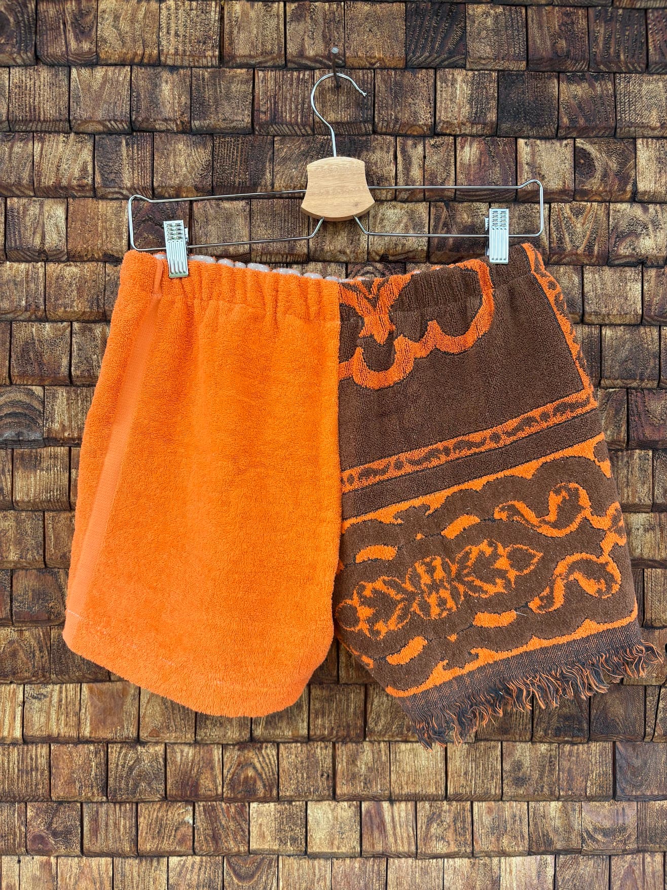 Rückansicht der Upcycling-Frotteeshorts „Dark Sunset ‘74“ von Old Wave auf einem Kleiderbügel vor einer Holzwand. Die Rückseite führt das asymmetrische Farb- und Musterkonzept fort: Die linke Gesäßhälfte ist komplett aus schlichtem, orangem Frotteestoff gefertigt, während das rechte Hosenbein das ausdrucksstarke, braun-orangefarbene Retro-Fliesen- und Ornamentmuster zeigt.