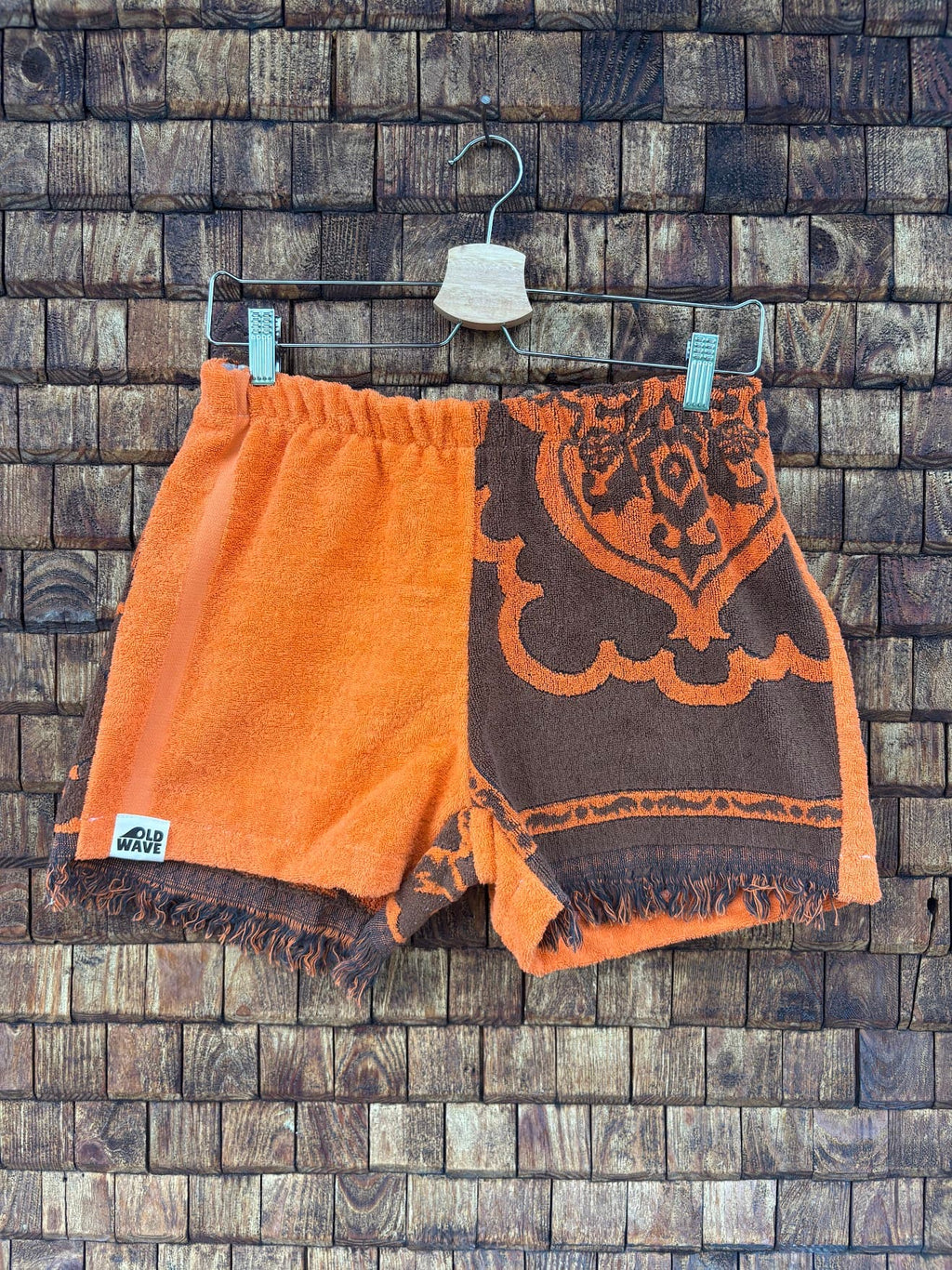 Vorderansicht der Upcycling-Frotteeshorts „Dark Sunset ‘74“ von Old Wave auf einem Kleiderbügel vor einer rustikalen Holzwand. Das kontrastreiche 70er-Jahre-Design besticht durch eine klare Zweiteilung: Das rechte Hosenbein besteht aus unifarbenem, leuchtend orangefarbenem Frottee mit dem Logo-Patch am Saum. Das linke Hosenbein zeigt ein großes, dunkelbraunes Paisley- und Ornamentmuster auf orangefarbenem Grund, abgerundet durch einen gefransten Saum.
