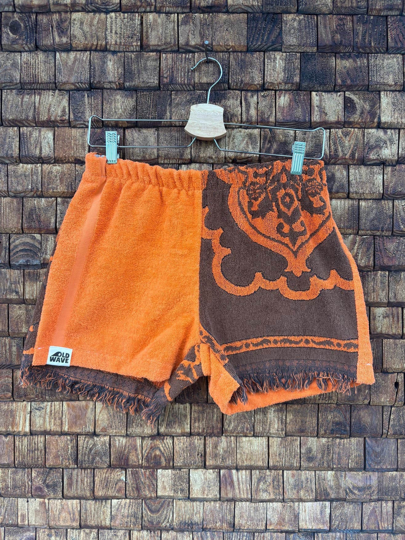 Vorderansicht der Upcycling-Frotteeshorts „Dark Sunset ‘74“ von Old Wave auf einem Kleiderbügel vor einer rustikalen Holzwand. Das kontrastreiche 70er-Jahre-Design besticht durch eine klare Zweiteilung: Das rechte Hosenbein besteht aus unifarbenem, leuchtend orangefarbenem Frottee mit dem Logo-Patch am Saum. Das linke Hosenbein zeigt ein großes, dunkelbraunes Paisley- und Ornamentmuster auf orangefarbenem Grund, abgerundet durch einen gefransten Saum.