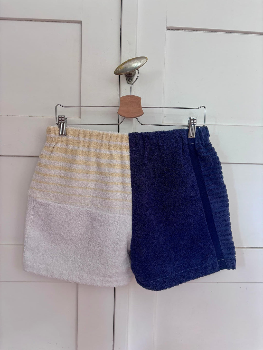Detailansicht der „Deep Coast 68“ Frottee-Shorts von Old Wave, aufgehängt an einem Holzkleiderbügel. Diese Seite zeigt ein dreigeteiltes Patchwork-Design aus upgecycelten Handtüchern: Ein dunkelblaues Element oben links, darunter weiß-gelbe Streifen, gepaart mit einer rechten Hosenbeinseite aus weißem Stoff mit hellblauen, geschwungenen Mustern.