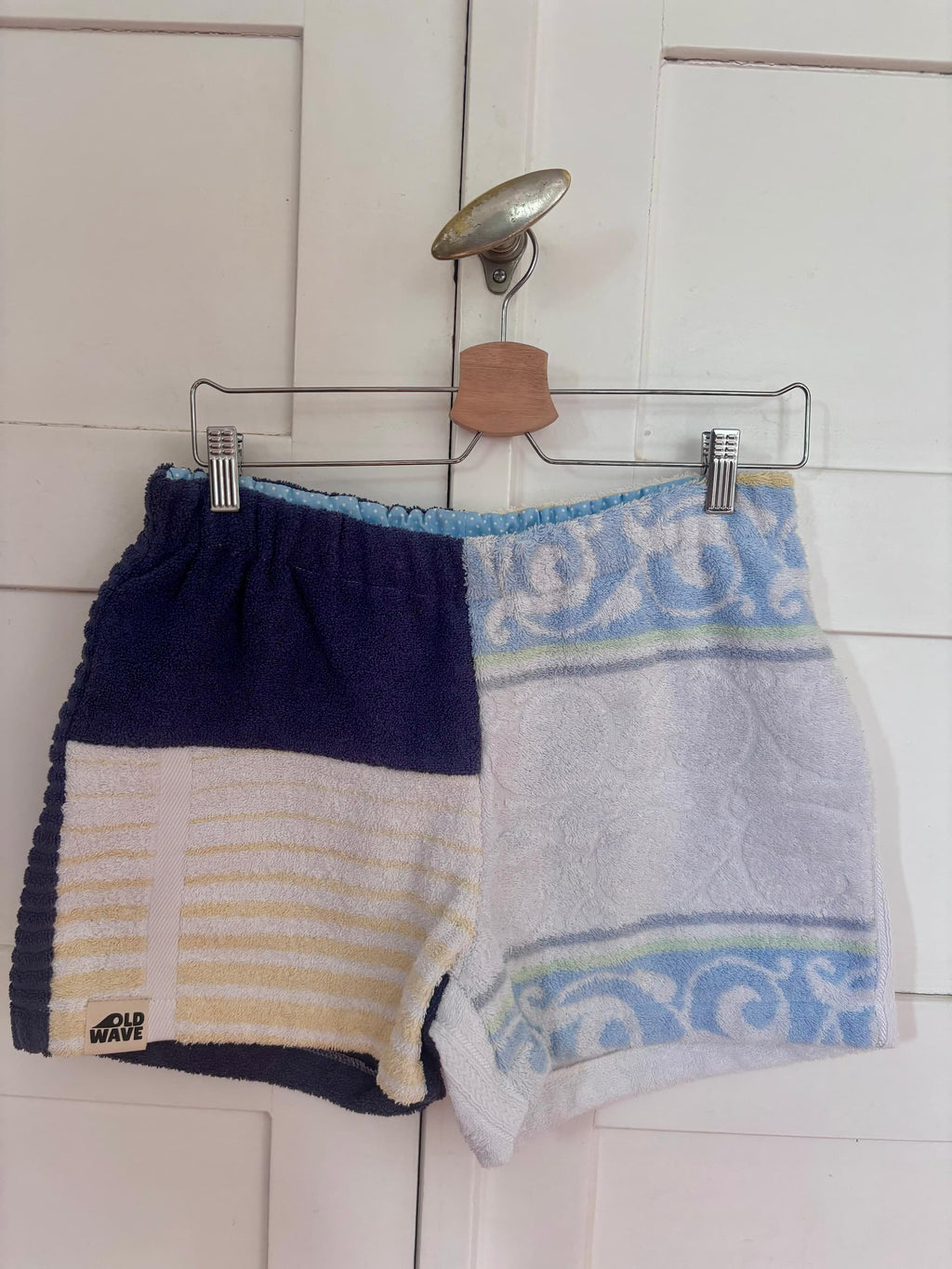 Upcycling-Shorts „Deep Coast 68“ von Old Wave auf einem Kleiderbügel vor einer weißen Holztür. Das Design zeigt eine geteilte Optik: Die linke Hosenbeinseite besteht aus weiß-gelb gestreiftem und rein weißem Frottee-Handtuchstoff, das rechte Hosenbein ist komplett in Dunkelblau gehalten.