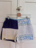 Upcycling-Shorts „Deep Coast 68“ von Old Wave auf einem Kleiderbügel vor einer weißen Holztür. Das Design zeigt eine geteilte Optik: Die linke Hosenbeinseite besteht aus weiß-gelb gestreiftem und rein weißem Frottee-Handtuchstoff, das rechte Hosenbein ist komplett in Dunkelblau gehalten.