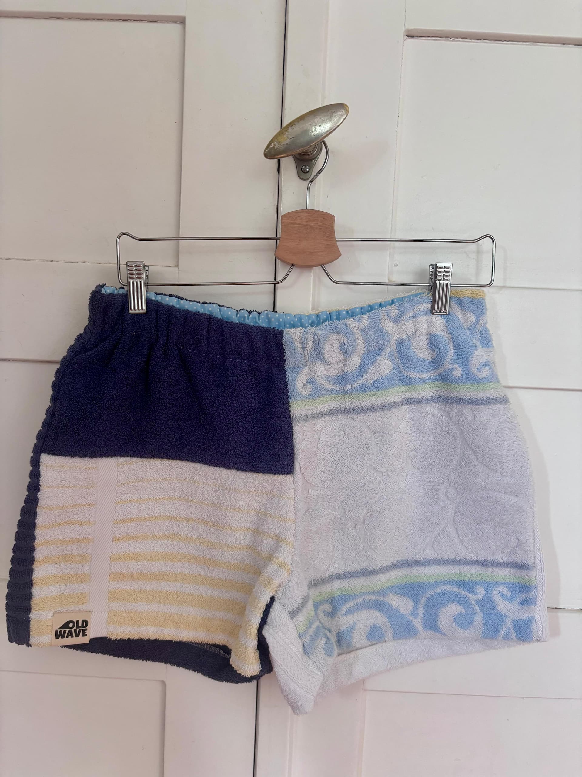 Upcycling-Shorts „Deep Coast 68“ von Old Wave auf einem Kleiderbügel vor einer weißen Holztür. Das Design zeigt eine geteilte Optik: Die linke Hosenbeinseite besteht aus weiß-gelb gestreiftem und rein weißem Frottee-Handtuchstoff, das rechte Hosenbein ist komplett in Dunkelblau gehalten.