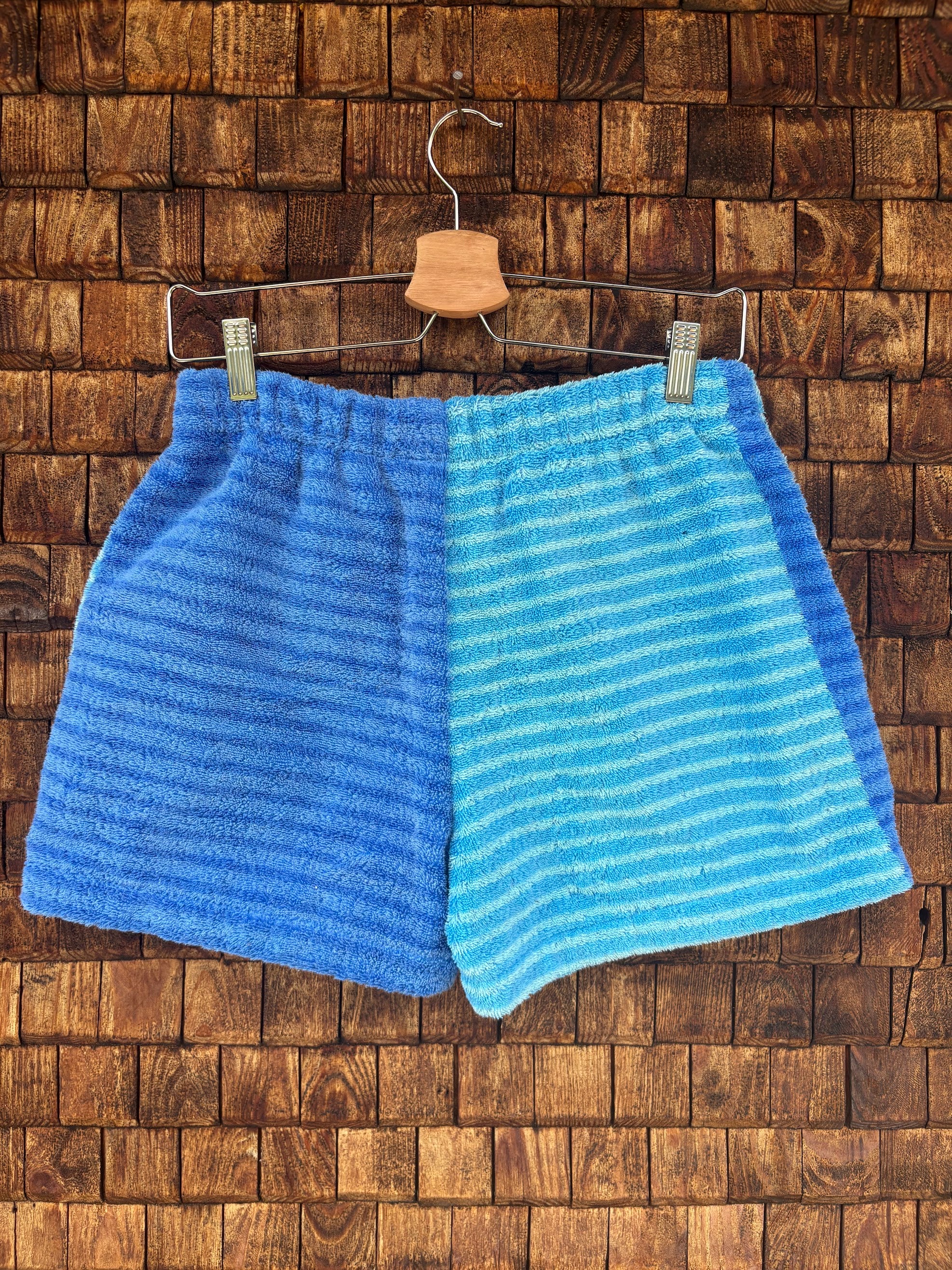 Rückansicht der Upcycling-Frotteeshorts „Deep Ocean ‘88“ von Old Wave auf einem Kleiderbügel vor einer Holzwand. Die Rückseite präsentiert ein harmonisches, hälftig geteiltes Color-Blocking: Die linke Gesäßhälfte besteht komplett aus dem dunkelblau gestreiften Frotteestoff, während die rechte Hälfte durchgehend im hellblau-türkis gestreiften Retro-Muster gehalten ist.