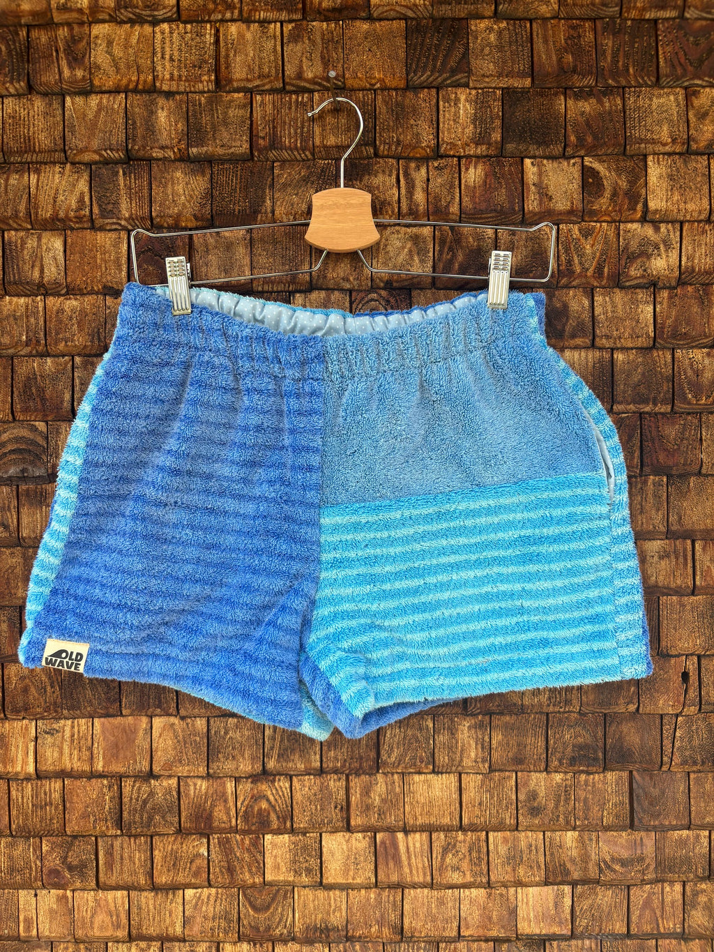 Vorderansicht der Upcycling-Frotteeshorts „Deep Ocean ‘88“ von Old Wave auf einem Kleiderbügel vor einer rustikalen Holzwand. Das maritime Design in Blautönen zeigt ein linkes Hosenbein aus dunkelblau gestreiftem Frottee mit dem Old Wave-Logo am Saum. Das rechte Hosenbein kombiniert eine obere Hälfte in meliertem Jeansblau mit einer untere Hälfte aus hellblau-türkis gestreiftem Vintage-Frotteestoff.