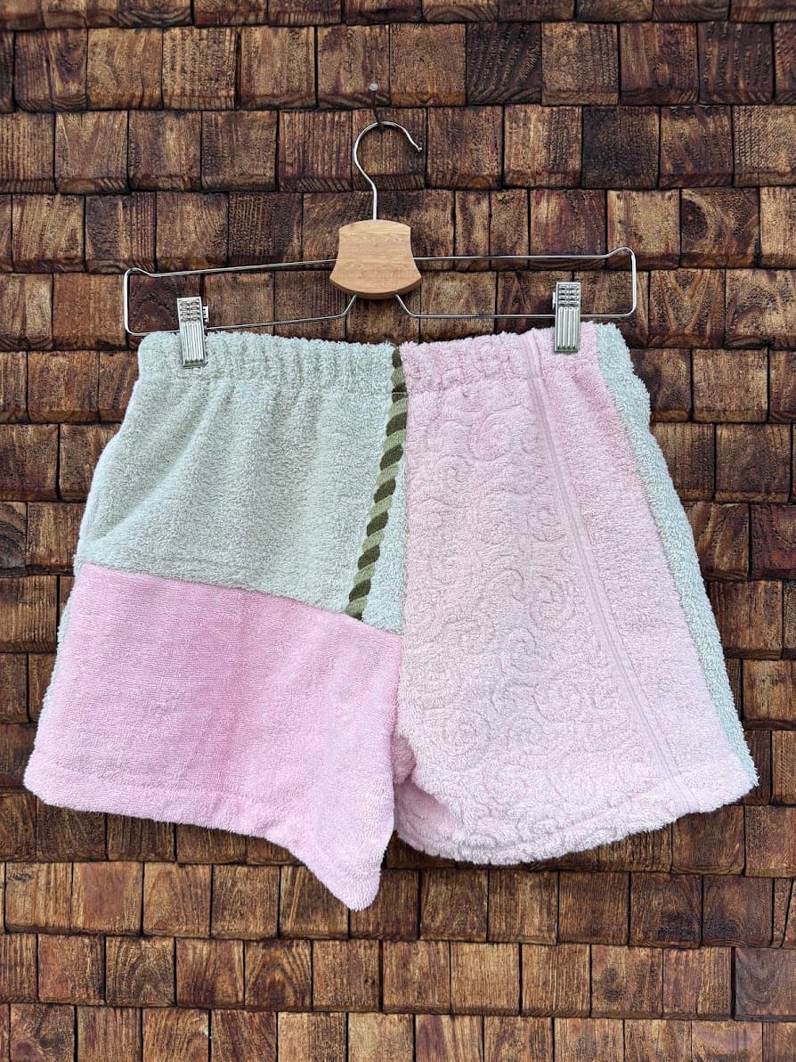 Rückansicht der Upcycling-Frotteeshorts „Dusty Resort ‘87“ von Old Wave auf einem Kleiderbügel vor einer Holzwand. Die obere Hälfte der Shorts inklusive des elastischen Bunds ist in Lachsrosa gehalten, während die untere Hälfte farblich zweigeteilt ist in Hellgrau links und Weiß rechts. Ein modischer Akzent wird durch eine aufgesetzte Gesäßtasche aus gemustertem Vintage-Bortenstoff sowie eine dekorative Lasche auf der linken Seite gesetzt.